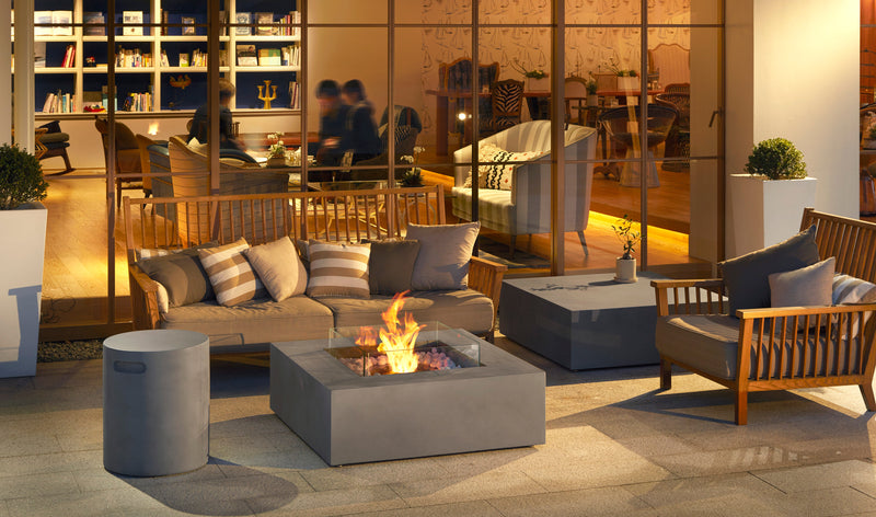 A cozy outdoor seating area with a fire pit, wooden furniture, and cushions, in front of a blurred indoor space with people.